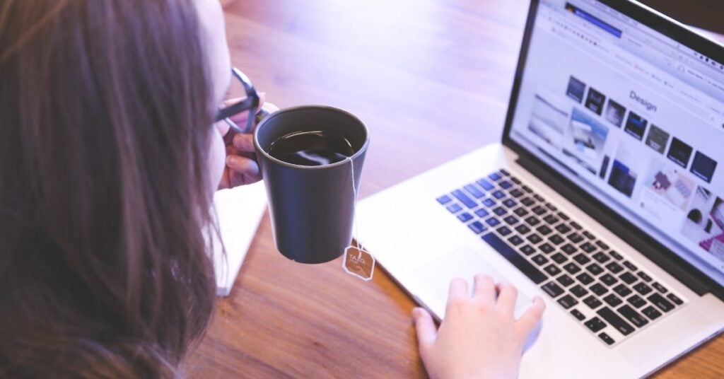 Woman with coffee in one hand while using a laptop.